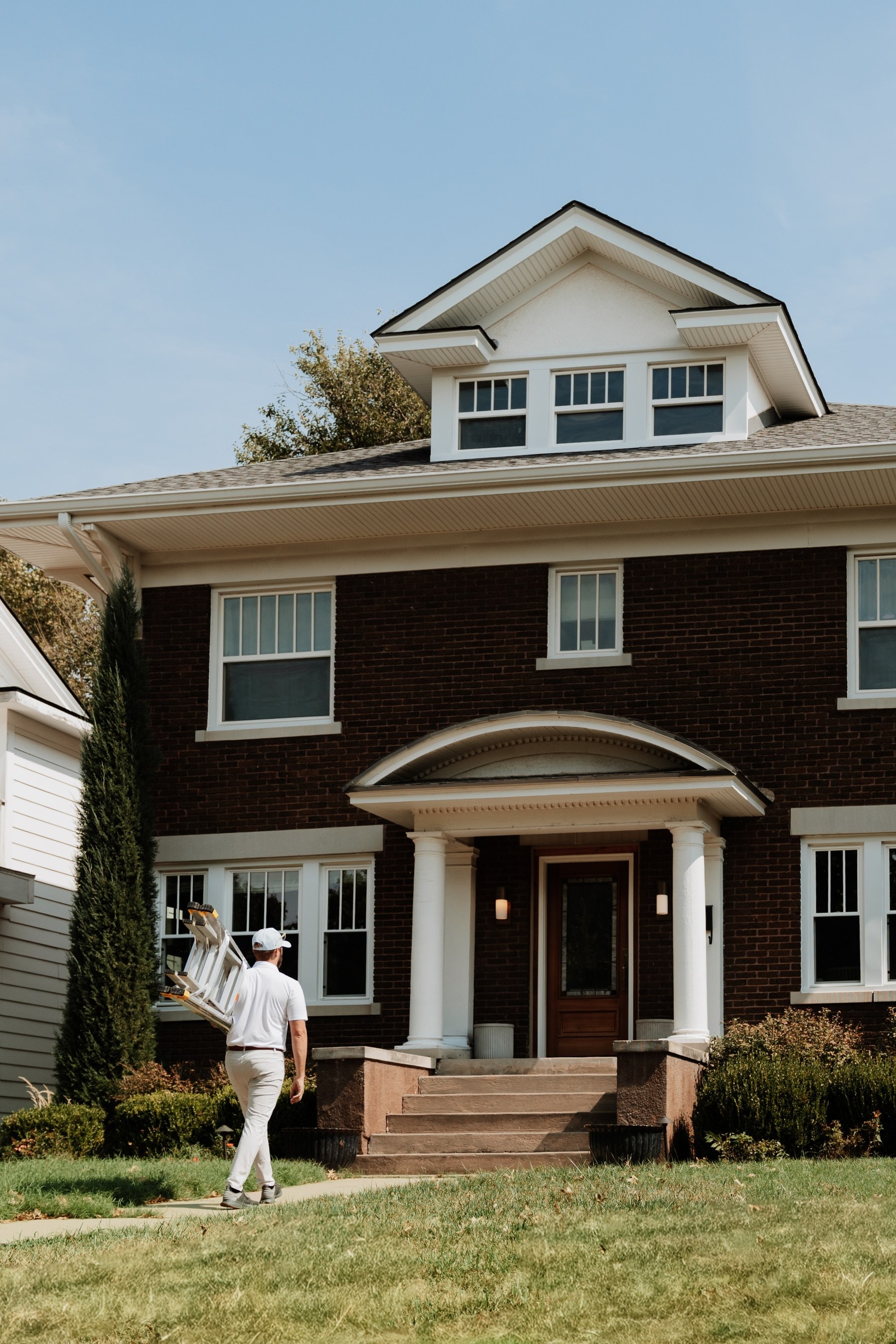 A Brella technician carrying a ladder up the front walk of a brick two-story home