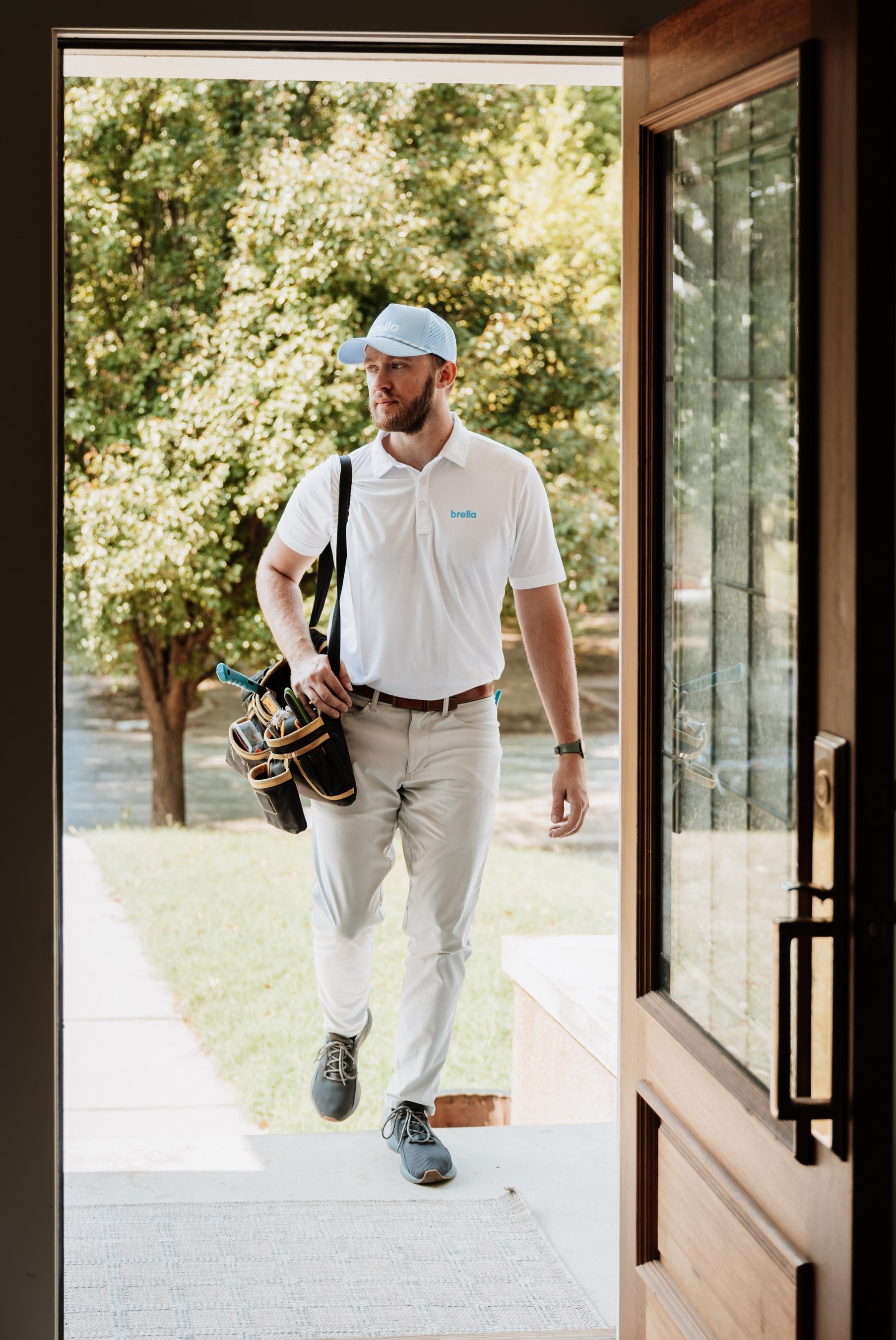 A Brella technician in a white polo and blue cap walking in through the front door with a tool bag